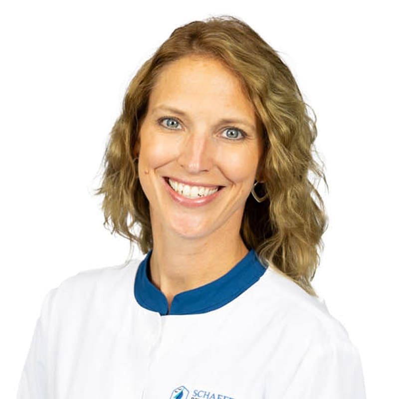 A smiling woman dentist in a white coat with blue collar, posing against a white background at an East Lansing, MI dental clinic.