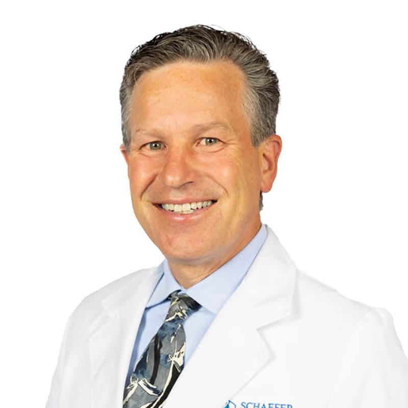 Smiling middle-aged East Lansing, Michigan dentist with short gray hair in a white coat, blue shirt, and tie against white backdrop.