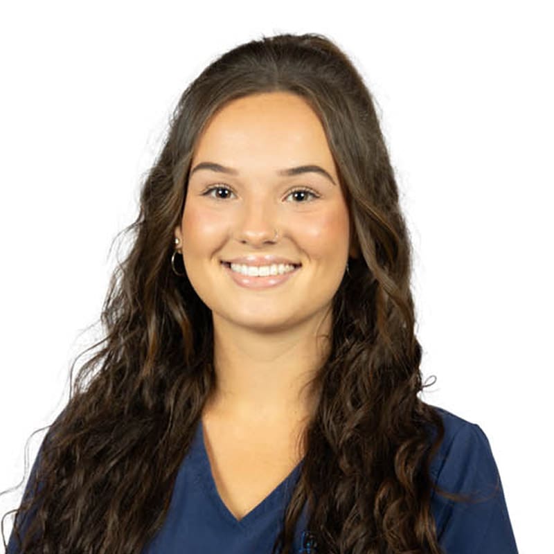 A confident young woman with long, wavy brown hair smiles in a navy top, ideal for a dentist’s profile in East Lansing, Michigan.