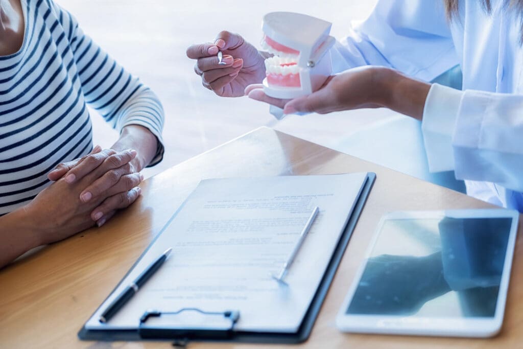 Dentist in East Lansing demonstrates dental implant procedure using a model to a seated patient with treatment paperwork and tablet.