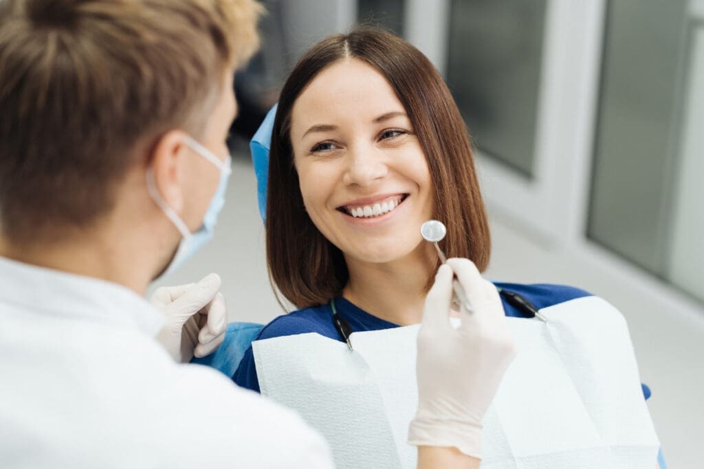 Female patient in dental chair smiles as dentist in PPE uses mirror, demonstrating standard patient care at East Lansing clinic.