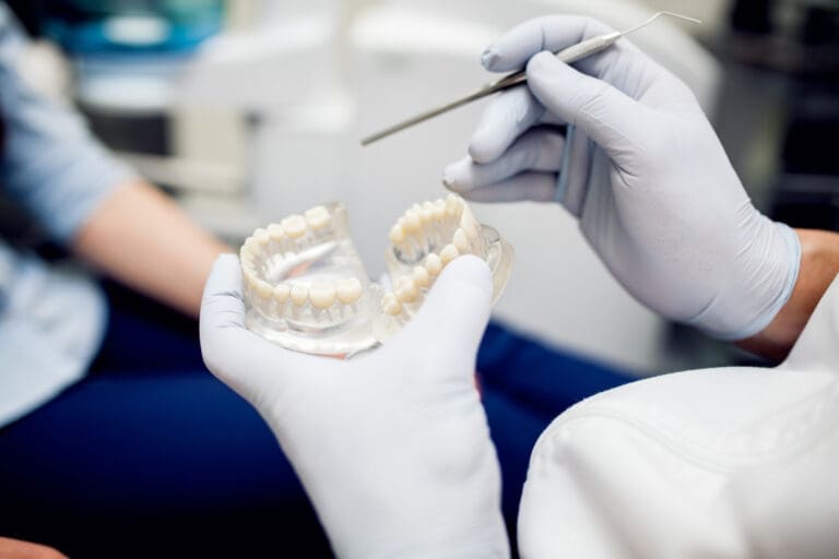 Dentist demonstrating prosthetic teeth with a dental mold and instrument at East Lansing clinic, patient observing in background.