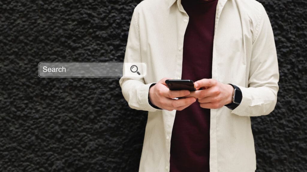 A person stands by a black wall in casual attire, holding a phone with a digital search bar—ideal for East Lansing dental searches.